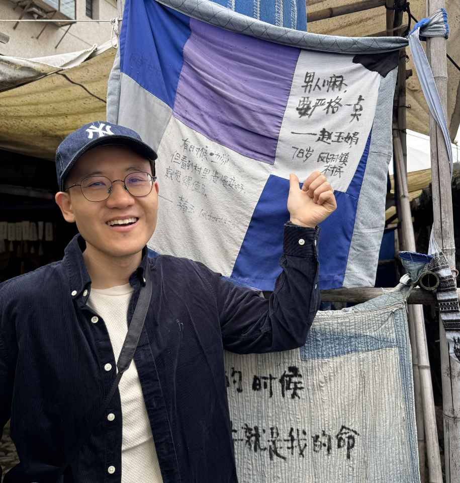 男生戴着鸭舌帽和眼镜,在写满手写字的拼布幡前微笑着竖起拇指