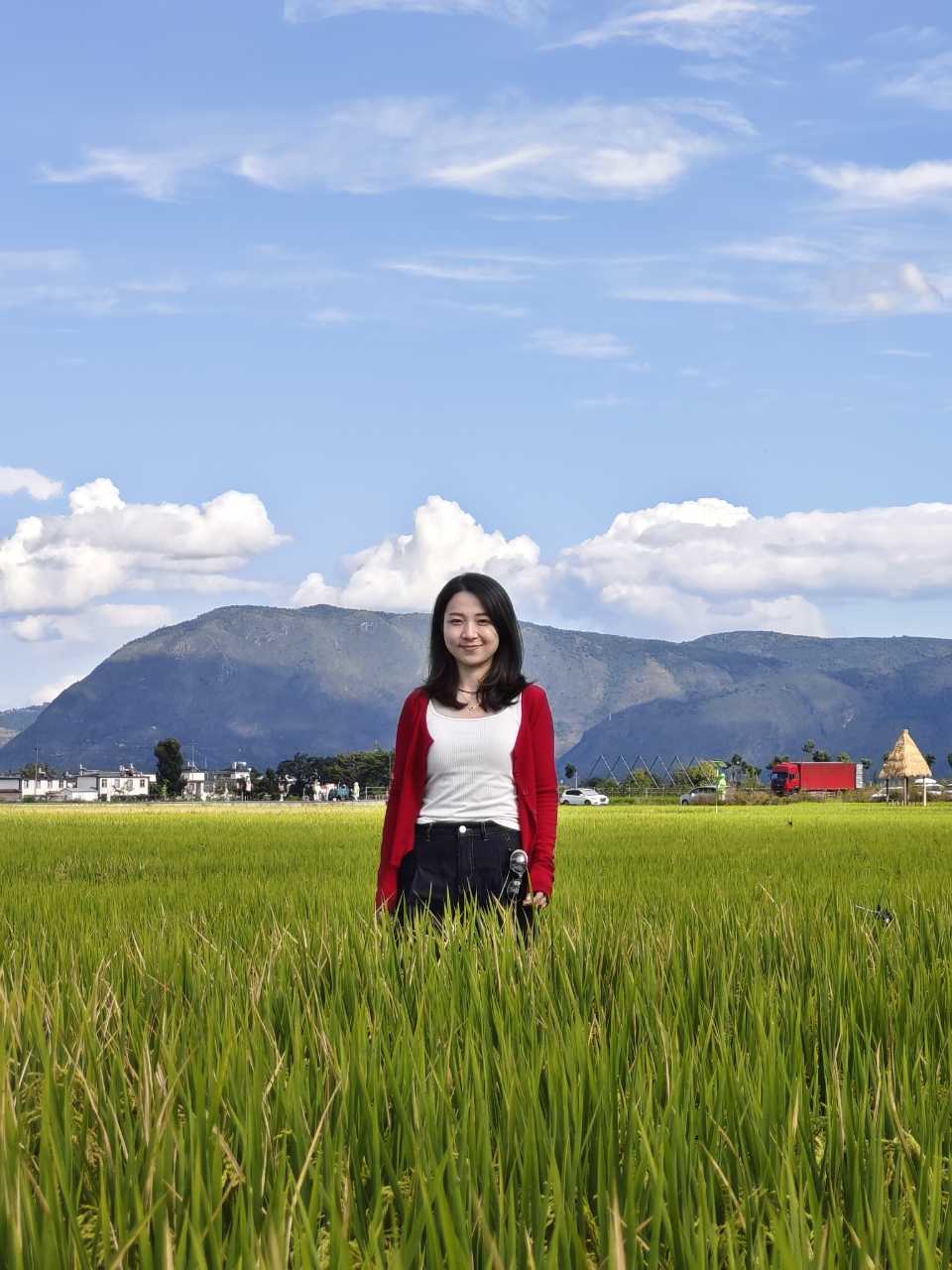 女生站在稻田中央微笑着看向镜头,身后是苍山与大片云朵